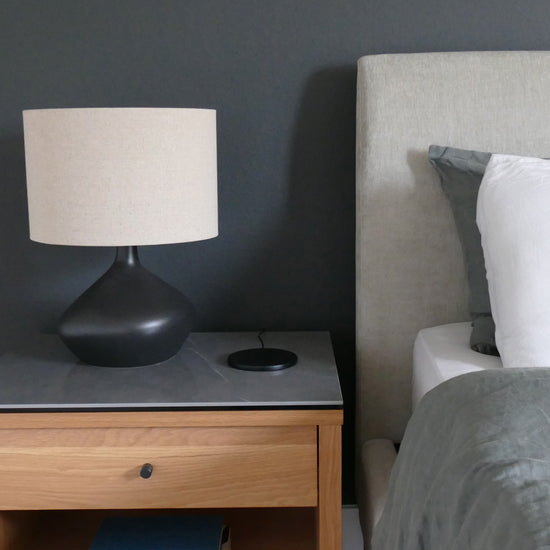 Left: Modern black and beige linen lamp on wooden dresser with marble top. Moonstone black charger cover case on wireless charger. Right: Gray upholstered bed with sage green and bright white bedding. 